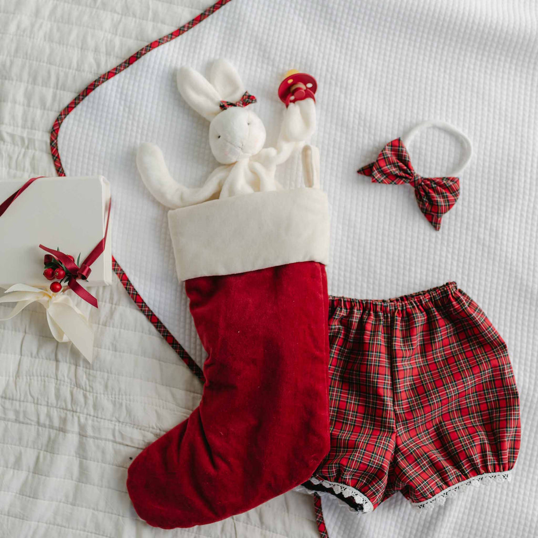 A Christmas stocking in red features the Gabriel Silly Bunny Buddy, which doubles as a pacifier holder for baby-safe use, sitting on the bed. Next to it lies a set of red plaid shorts accompanied by a matching bow tie and headband. Nearby rests a white gift box, elegantly tied with a red ribbon and decorated with a holly sprig.