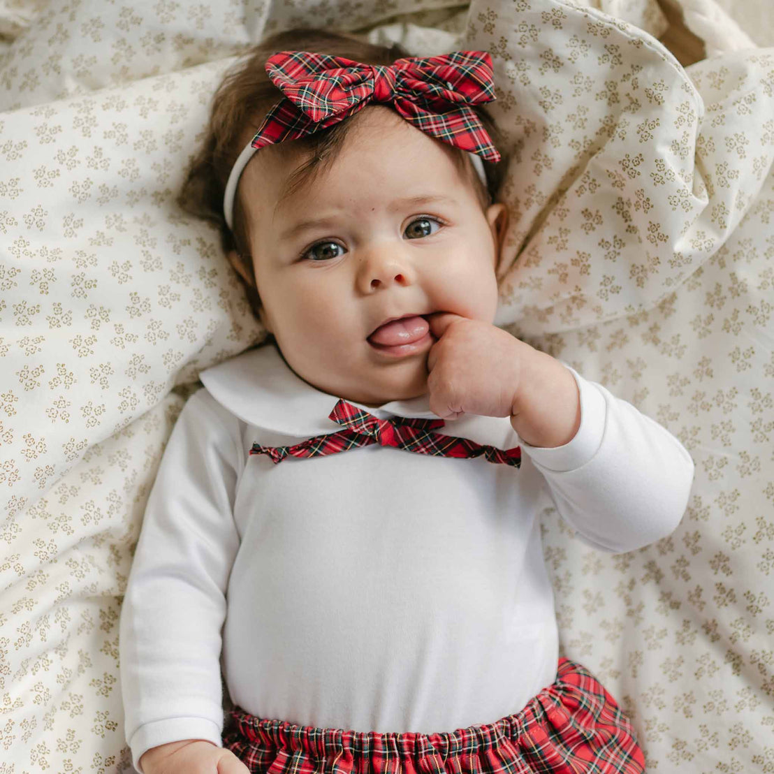 A baby lies on a patterned blanket, wearing the Gabriella Bloomer Set with red plaid accents and matching headband, sucking on one finger while looking up, perfectly dressed in their holiday outfit.