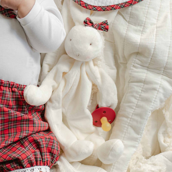 A baby in a red plaid outfit lies next to the Gabriel Silly Bunny Buddy, a white stuffed toy with a plaid bow, securely holding a pacifier with its safe clip. The cozy scene unfolds on the backdrop of a soft, cream-colored blanket.