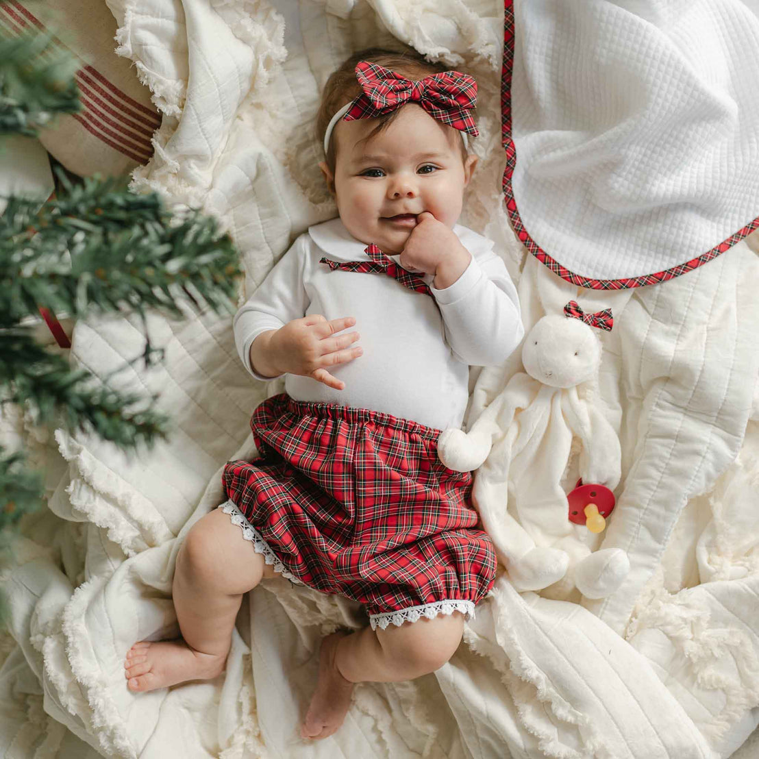 A baby dressed in the Gabriella Bloomer Set—a cozy holiday outfit featuring a white cotton onesie with red plaid bloomers and a matching headband—rests on a soft blanket. The little one cuddles a white plush rabbit, surrounded by plush toys and greenery, creating an adorable festive scene.