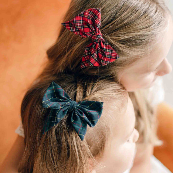 Two girls with plaid bow hair clips in their hair sit together. The girl in front flaunts a handmade red plaid bow, while the one behind sports a dark blue hair clip. They face partially away, focusing on something outside the frame. The background is softly blurred, adding a touch of mystery.