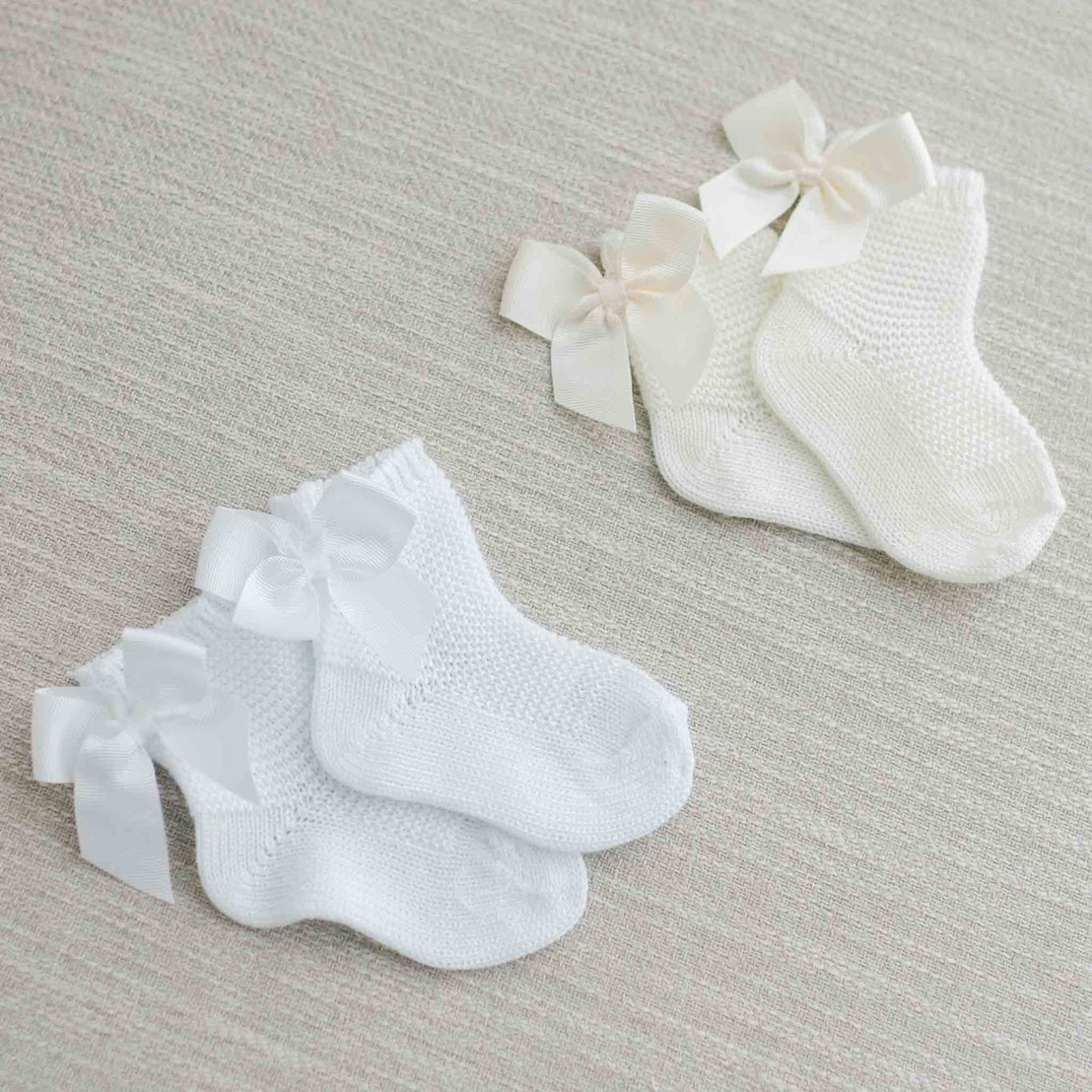 Two pairs of Garter Stitch Socks with Bow, one in white and the other in ivory, presented on a soft beige fabric background.