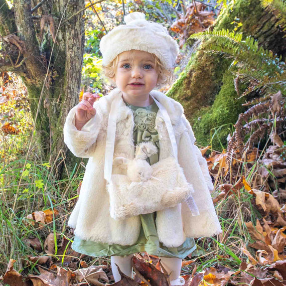 A young child in the charming Glad Dreams Coat Set stands amidst fallen leaves and green grass in a forested area, holding a small object. Ferns and tree trunks surround the child, creating a natural, autumnal setting—an unforgettable unique baby gift moment.