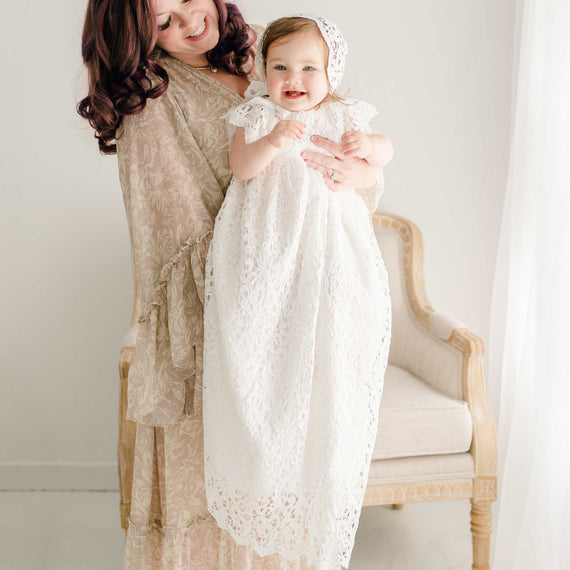 A mother holding her smiling baby daughter dressed in the Christening Gown and Bonnet. The mother is standing on front of a linen upholstered chair and smiling down at her daughter. 