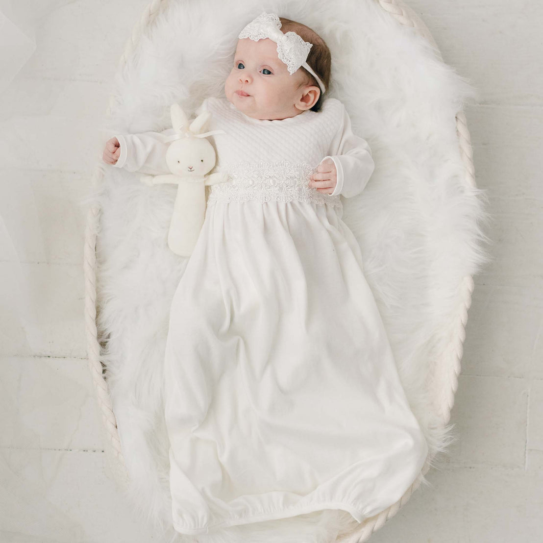 A baby with a headband lies in an upscale, cozy white basket, holding a stuffed rabbit, dressed in a Madeline Newborn Gown, on a fluffy rug.