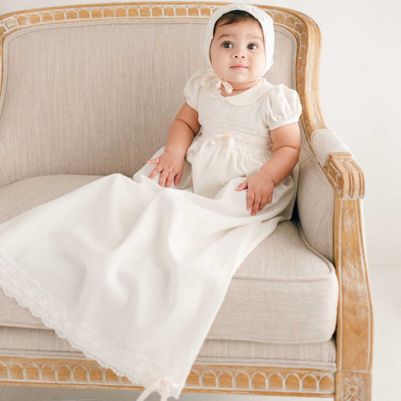 A newborn in an Emma Convertible Skirt & Romper Set sitting on an antique sofa, looking up with wide eyes. The baby's outfit flows down.