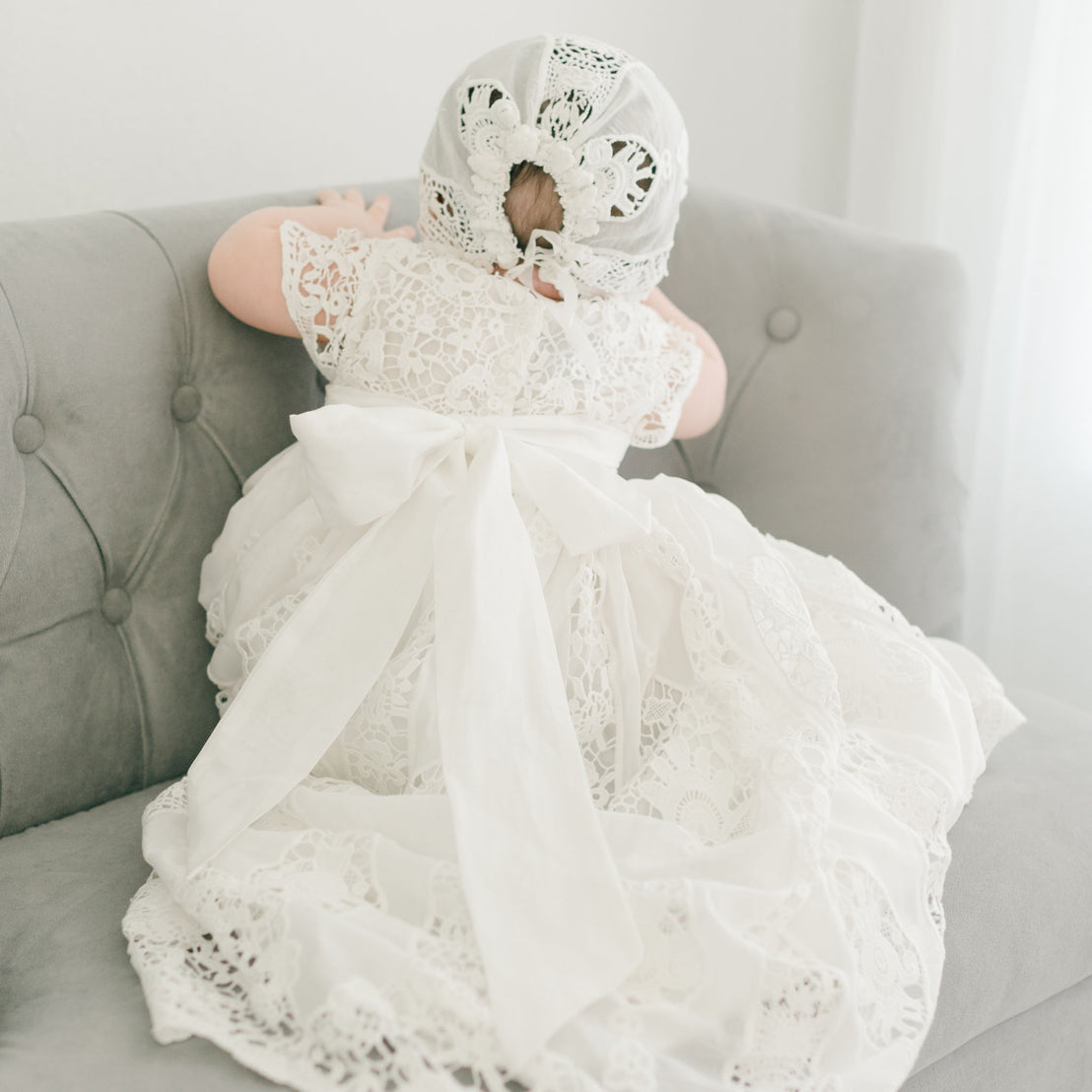 A baby wearing the Grace Lace Bonnet, adorned with a large white bow, sits on a soft, light gray sofa in a delicate ivory lace dress. The scene is illuminated with natural light filtering in.