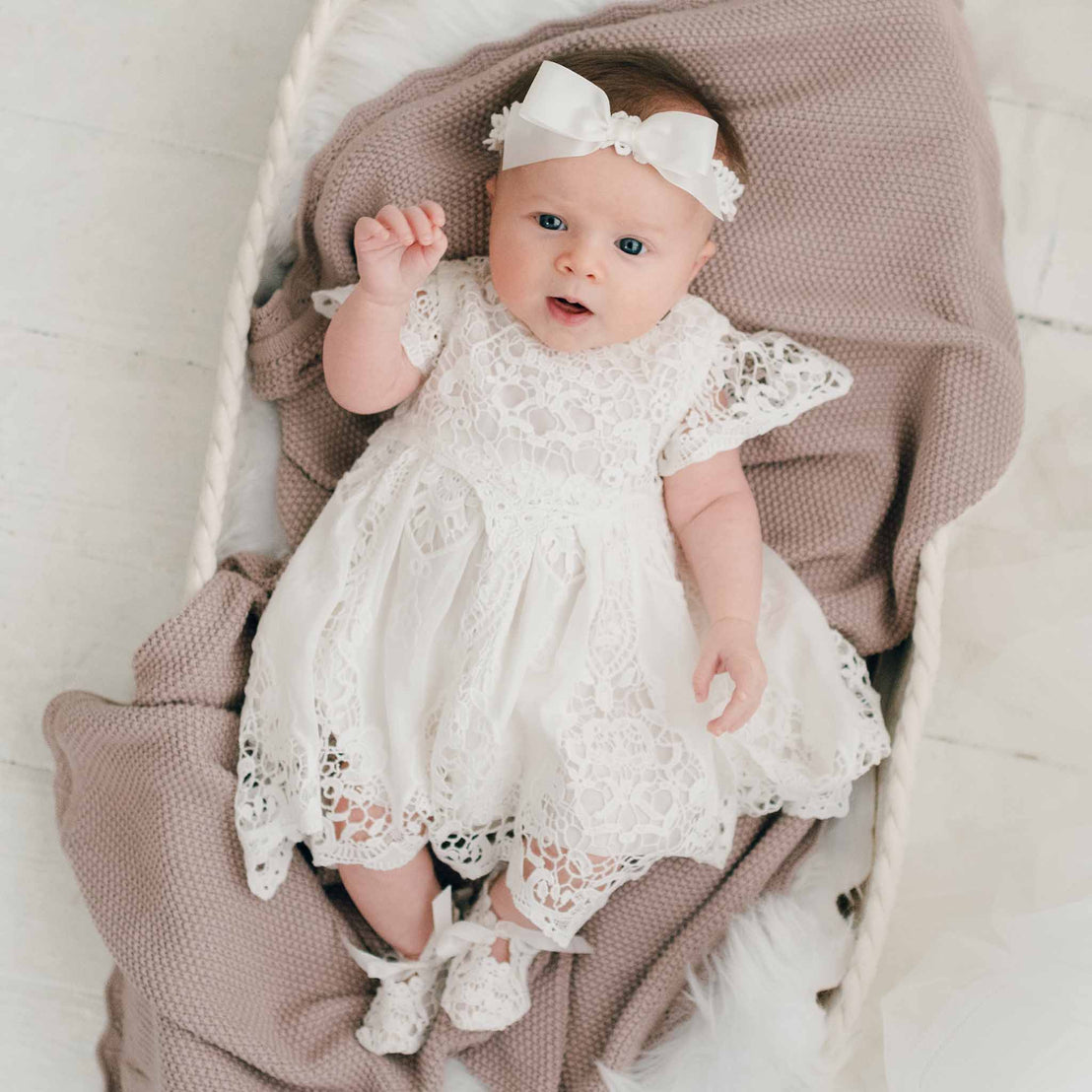 A baby girl in a white Grace Dress and headband lies in a cozy basket, looking up with a soft smile, surrounded by a knitted pink blanket.