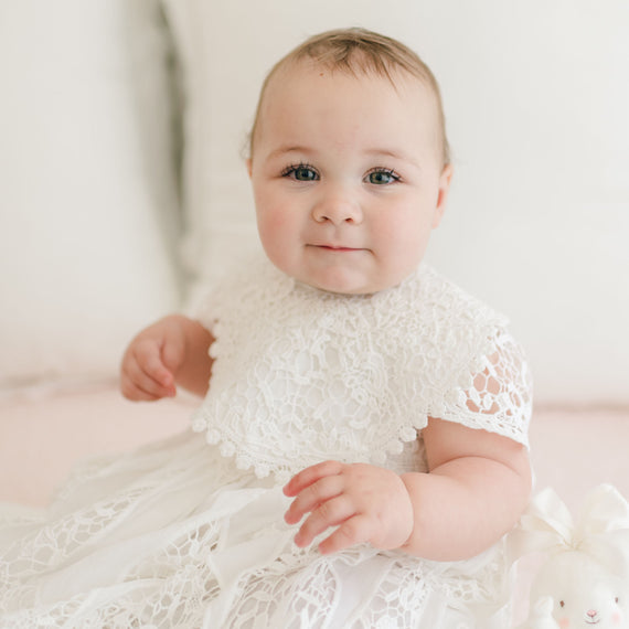 A baby with light skin and rosy cheeks is sitting and wearing a delicate white cotton Grace Lace Bib, reminiscent of a christening gown. The baby has short, light brown hair and blue eyes, gazing towards the camera with a soft smile. There is a plush toy with a bow visible in the lower corner.