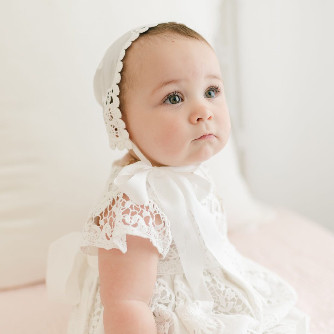 A baby with light skin and light-colored eyes gazes upward while seated on a soft surface. The infant is wearing the delicate, hand-crafted Grace Christening Gown & Bonnet, which is made of white lace and tied under the chin with a white ribbon. The softly blurred background enhances the gentle, serene mood.