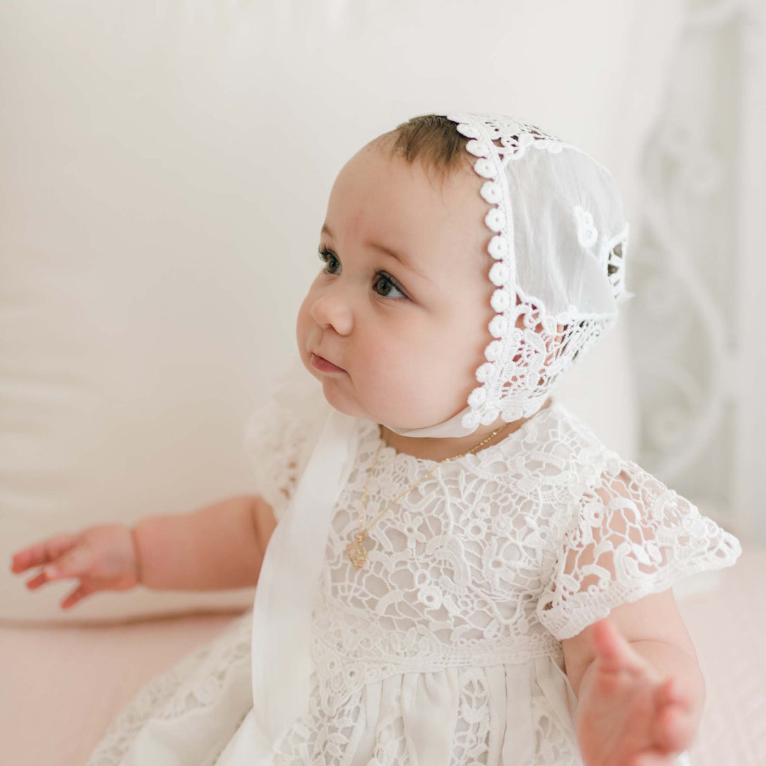 A baby wearing the Grace Dress, a delicate white lace outfit reminiscent of a flower girl dress, and a matching bonnet sits on a soft pink surface. The baby has a focused expression with arms extended outward, while the background is softly blurred with a light, neutral tone.