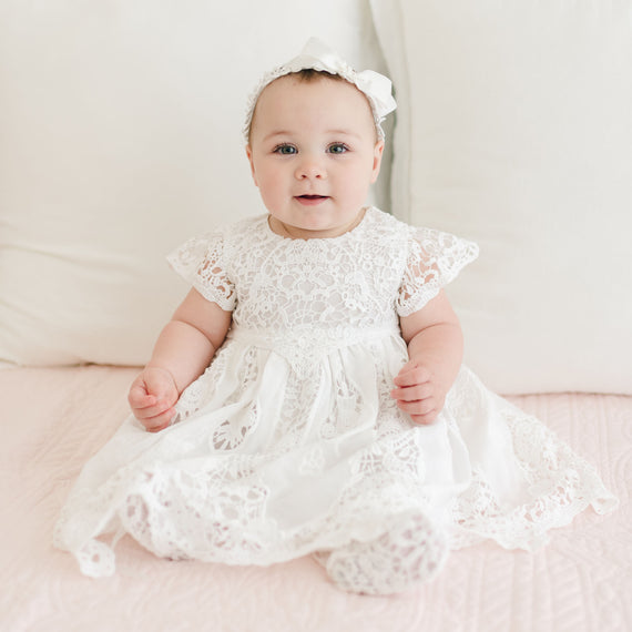 A baby wearing the Grace Dress, a white lace flower girl dress, and a matching headband with a bow sits on a light pink quilted surface, with white pillows in the background. The baby is smiling and has hands resting gently on the lap.