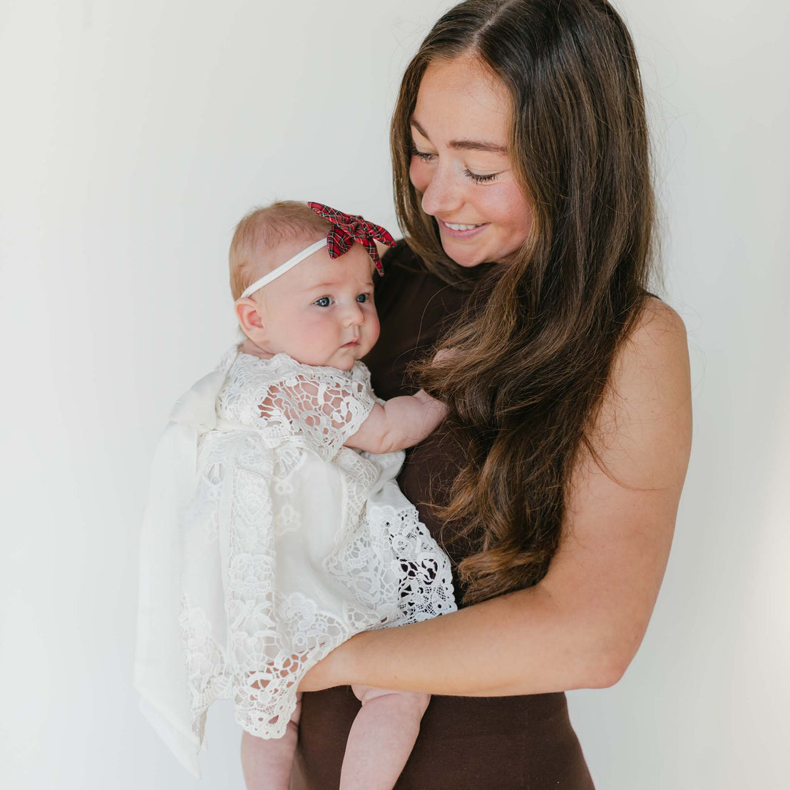 A woman with long brown hair smiles warmly as she cradles a baby dressed in a Grace Holiday Dress with a lace bodice. The baby, wearing a red headband and bow, enhances the festive Holiday Season atmosphere against the simple white backdrop.