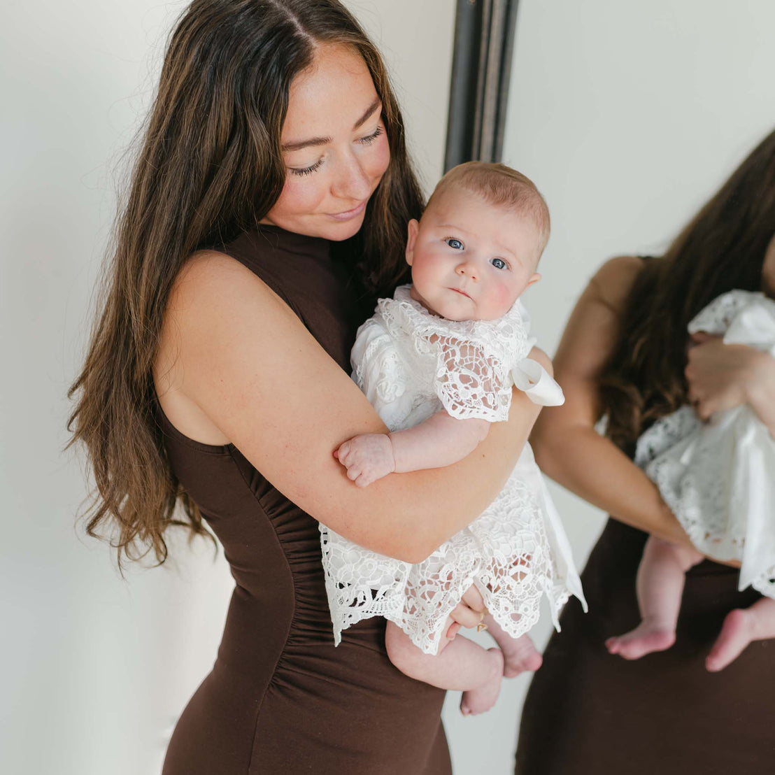 A woman with long brown hair stands by a mirror in her elegant Grace Holiday Dress, gently looking down at a baby dressed in a white lace outfit. It's a tender moment that captures the warmth of the season.