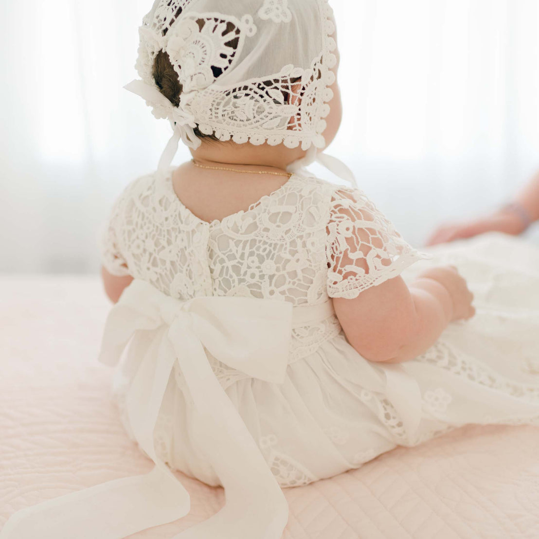 A baby wearing the Grace Christening Gown & Bonnet, made from a hand-crafted silk and cotton blend, sits facing away. The gown showcases a large white ribbon tied in a bow at the back. Soft lighting and a light background create an airy and gentle atmosphere.