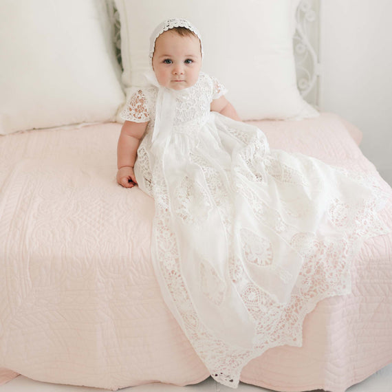 A baby wearing the Grace Christening Gown & Bonnet, crafted from a luxurious silk and cotton blend, sits on a bed adorned with a light pink quilt. The infant looks directly at the camera with a calm expression, framed by white pillows and an ornate white metal headboard in the background.