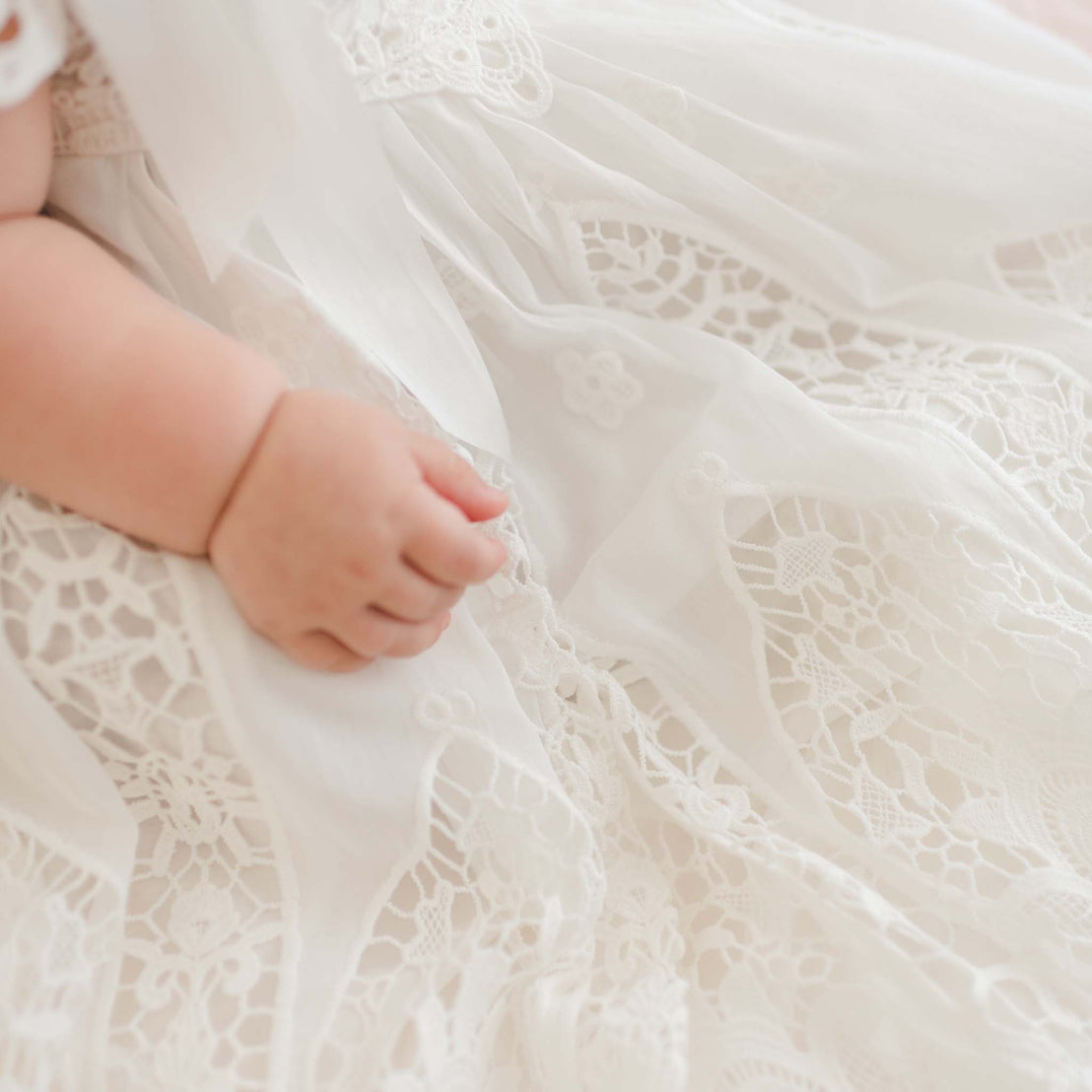 A close-up image of a baby dressed in the "Grace Christening Gown & Bonnet." The baby's hand, visible on the left side, rests on the delicate lace fabric, revealing intricate floral patterns. The soft and light silk and cotton blend material of the gown drapes gently, creating a serene and tender ambiance.