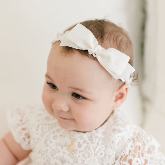 A baby wearing a white lace dress and the Grace Headband, adorned with a silk ribbon bow, looks off to the side with a slight smile. The baby's delicate features and soft expression create a serene and innocent ambiance in the photograph.