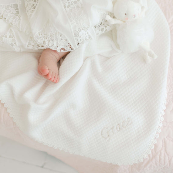 A close-up of a baby's feet peeking out from the Grace Personalized Blanket, adorned with lace trim and intricate embroidery. The name "Grace" is beautifully stitched onto the soft, white fabric. Beside the baby rests a white plush toy on the blanket. The scene exudes softness and delicacy, highlighted by light pastel tones that showcase an heirloom piece with a personalized touch.