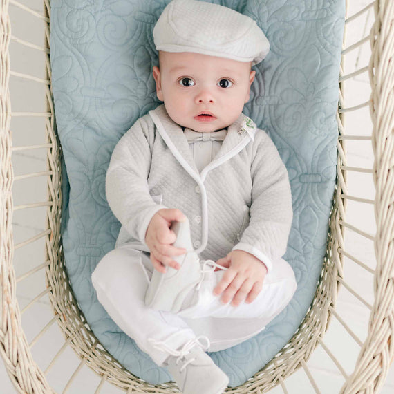 A baby dressed the Grayson 3-Piece Suit, a light gray outfit that includes the jacket, pants, and onesie, is lying in a blue quilted basket. The baby boy has a calm expression and is reaching out to touch one of his shoes.