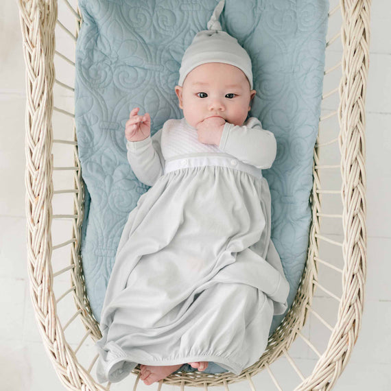 A baby is lying in a wicker bassinet decorated with a light blue quilted mattress. The baby is wearing the Grayson Newborn Layette and a matching hat with a knot on top. The baby has one hand near its mouth and looks up at the camera with a calm expression.