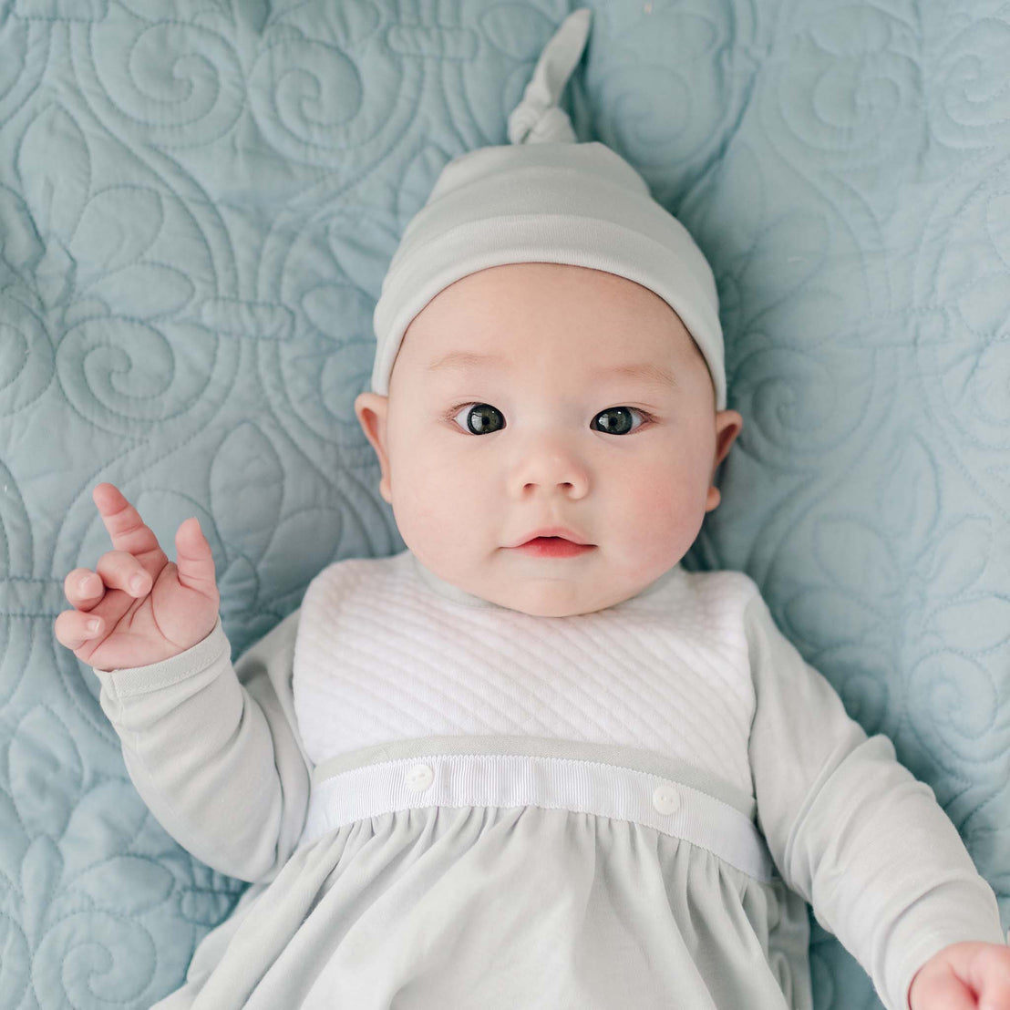 A baby lies on a light blue quilted blanket, dressed in an adorable Grayson Newborn Layette with a matching hat that has a small knot at the top. The baby has wide eyes and a slightly open mouth, looking directly at the camera with a small raised hand.