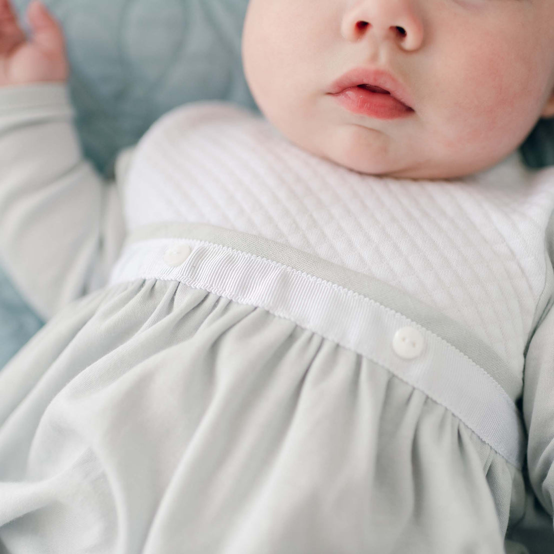 Close-up of a baby lying down, wearing a light-colored Grayson Newborn Layette with a quilted top and a gathered skirt. The focus is on the baby's lower face and torso, highlighting the soft fabric texture and buttons on the outfit. The background is softly blurred.