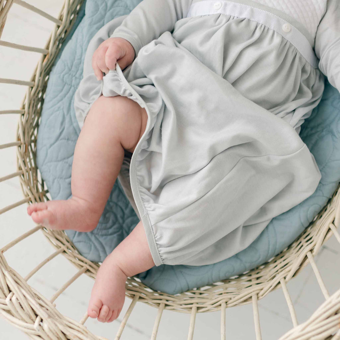 A baby dressed in a light blue Grayson Newborn Layette is lying on a pale blue quilted blanket in a woven bassinet. The baby is lifting the edge of their dress, revealing their legs and feet. The bassinet and surroundings create a soft, cozy atmosphere.