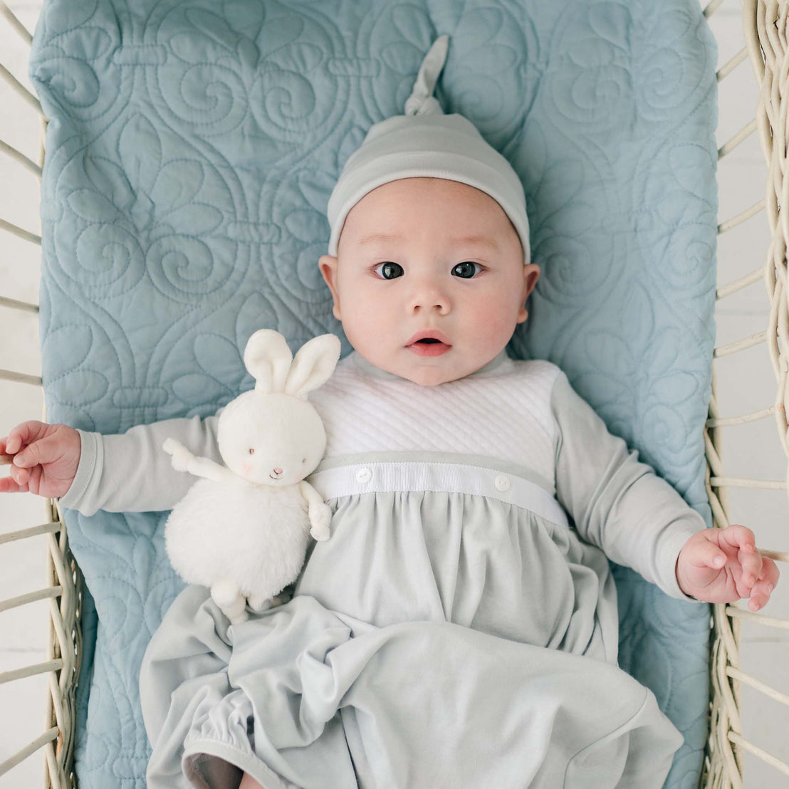 A baby lies in a crib on a quilted light blue blanket, wearing a matching light blue gown and hat. The Grayson Newborn Layette complements the serene setting as the baby holds a white plush bunny and looks up with wide eyes.