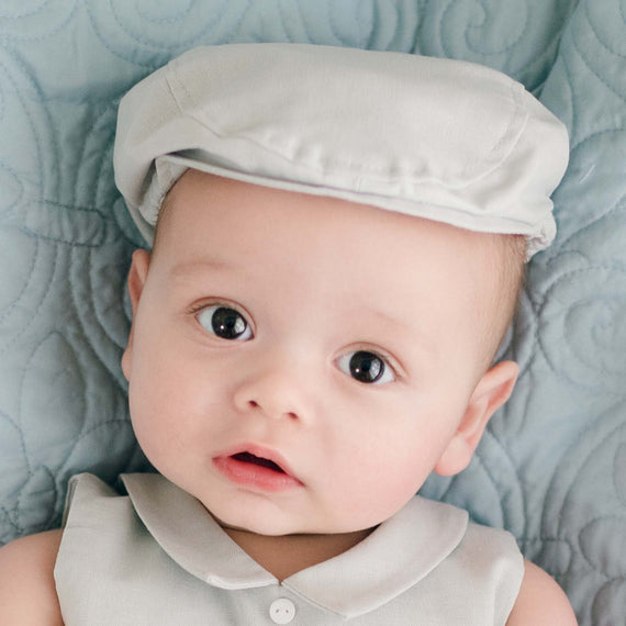 A baby with big, curious eyes is lying on a light blue quilted blanket. The baby is wearing the Grayson Linen Newsboy Cap with matching Linen Romper. The baby's expression is calm and slightly inquisitive.