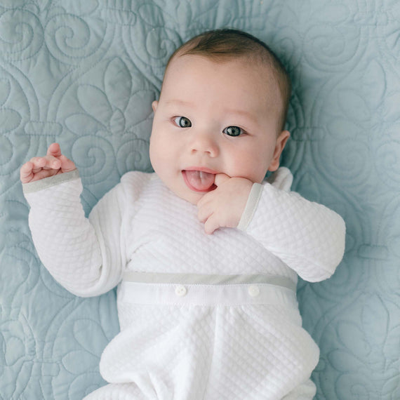 A baby wearing a designer take home outfit lies on a light blue quilted blanket. The newborn, dressed in a Grayson Quilted Romper, gazes at the camera with wide eyes, their tongue sticking out slightly as their right hand nears their mouth.