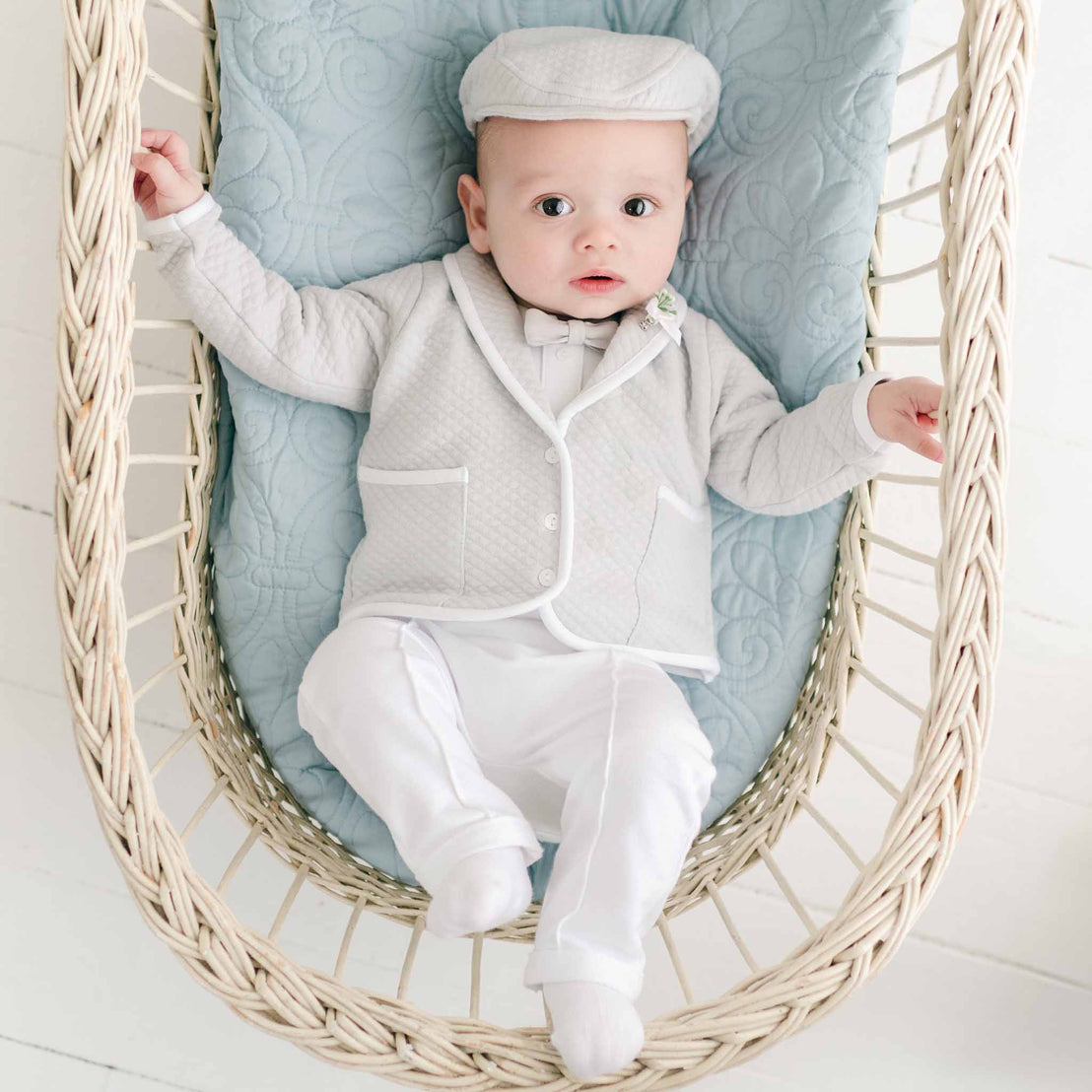 A baby dressed in the Grayson 3-Piece Suit, including the jacket, pants, and onesie, lies on a woven basket with a light blue quilted cushion. The baby has wide eyes and one hand resting on the side of the basket. The background is a light-colored, wooden floor.