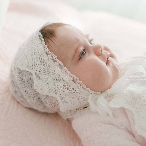 A baby lies on a soft pink blanket, wearing a delicate Hailey Knit Bonnet and light-colored clothing. The baby looks off to the side with a serene expression, showcasing their gentle features against a light and airy background while highlighting the charm of the Hailey Knit Bonnet.