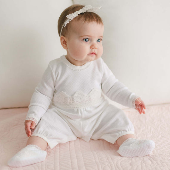 A baby wearing a Hailey Romper, white headband, and white socks is sitting on a pink quilted blanket. The baby has light-colored hair and blue eyes and is looking slightly to the side with a calm expression. The background is a plain white surface.