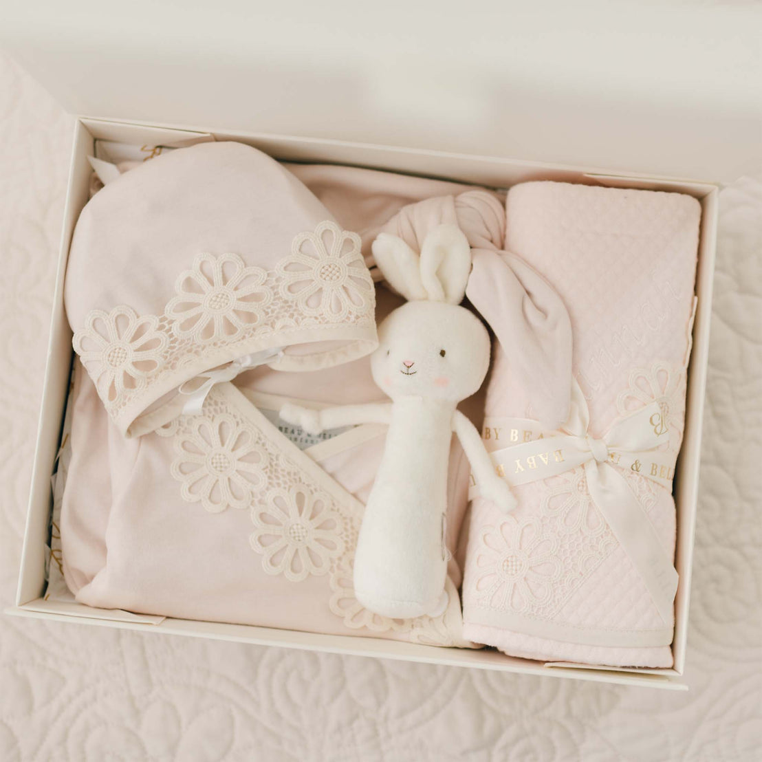 A neatly arranged Hannah Newborn Gift Set- Save 10% containing a pink newborn layette gown with daisy lace details, a matching hat, a soft 100% pima cotton blanket tied with a ribbon, and a white plush bunny toy. Handmade in the USA, the items are displayed on a white quilted surface.