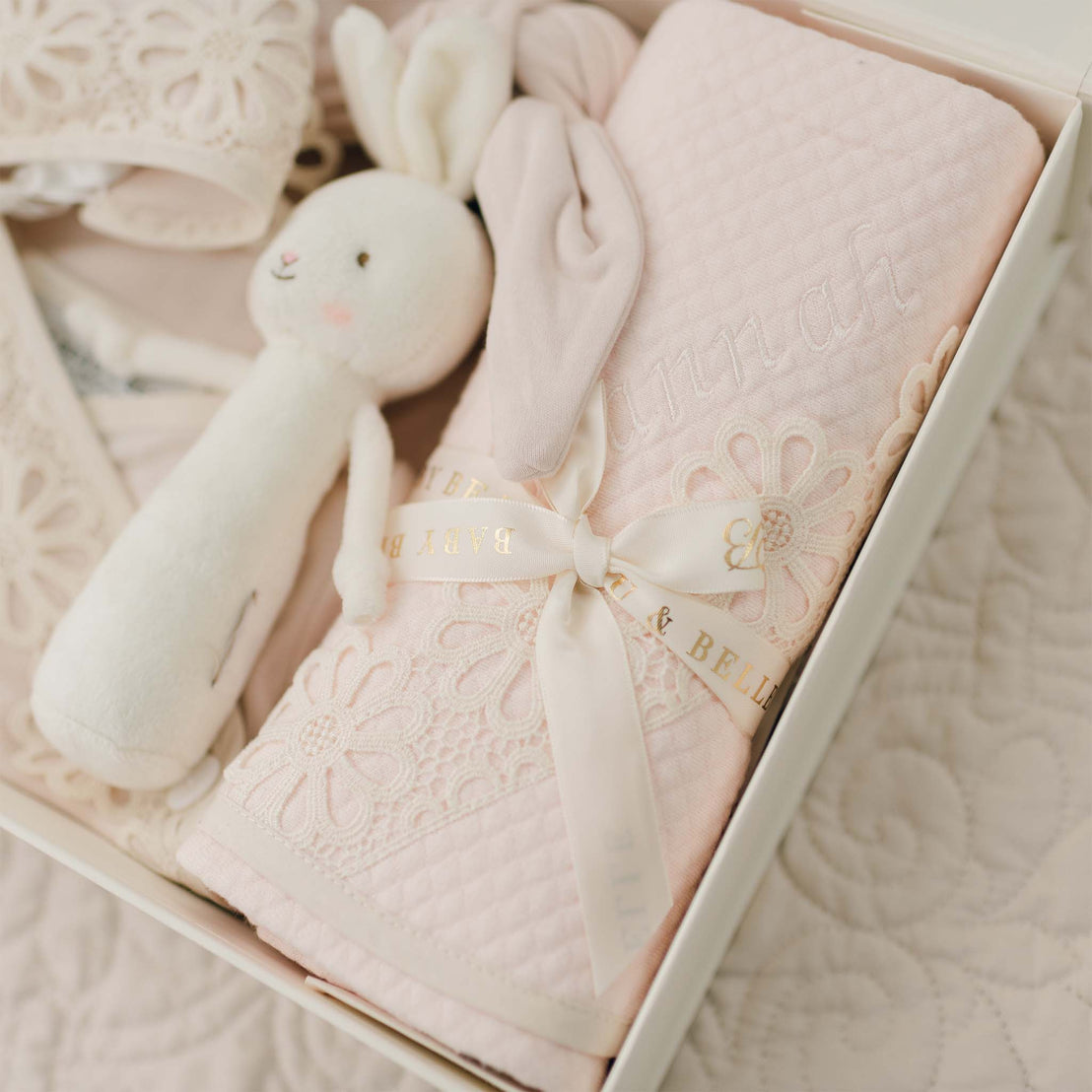 A gift box containing a soft, pink baby blanket adorned with embroidered flowers, a white plush bunny toy, and baby shoes. The 100% pima cotton blanket is tied with a ribbon featuring the text "Belle & Beau" and "BABY". The box is placed on a quilted surface. Introducing the **Hannah Newborn Gift Set- Save 10%**!