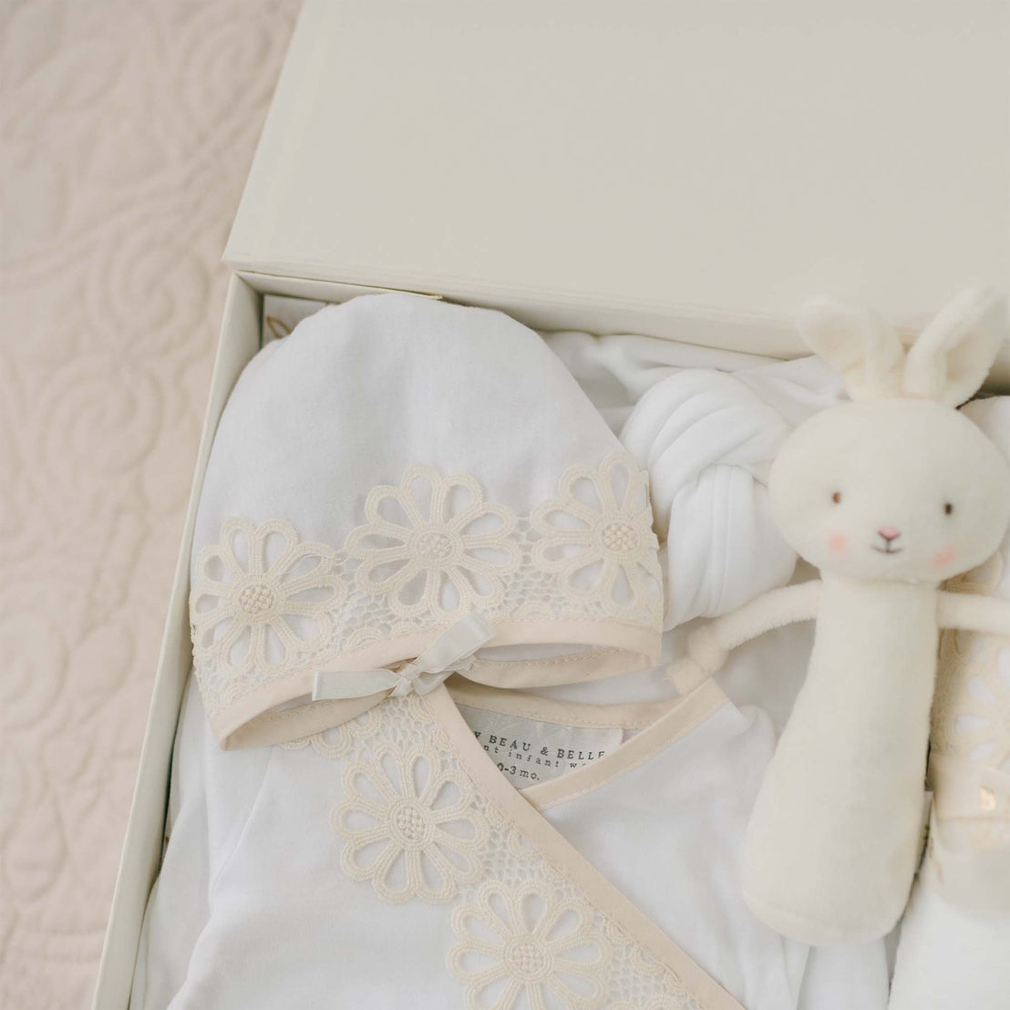 A neatly arranged Hannah Newborn Gift Set- Save 10% containing a white newborn layette gown adorned with delicate lace flower patterns, a matching hat, a knotted blanket made from 100% pima cotton, and a white plush bunny toy with subtle pink cheeks. All handmade in the USA, the items are elegantly placed against a soft, quilted background.