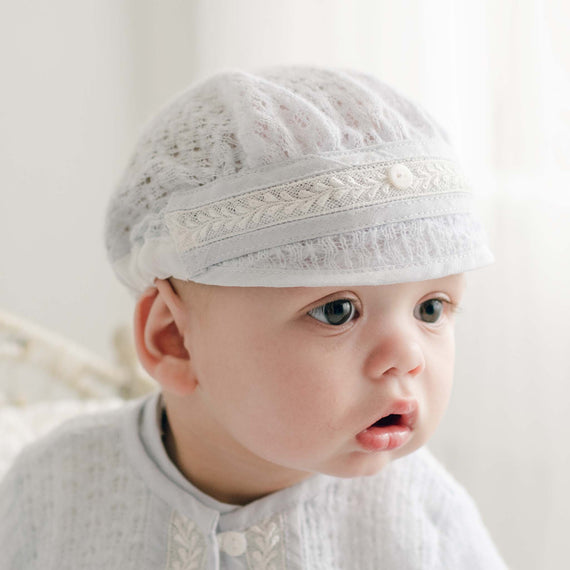 A newborn baby wearing a delicate Harrison Blue Knit Hat, handmade in the USA and featuring a small button detail, looks towards the right. The baby is dressed in a matching light-colored outfit. The softly lit background creates a gentle and serene atmosphere, ensuring the newborn baby's comfort.