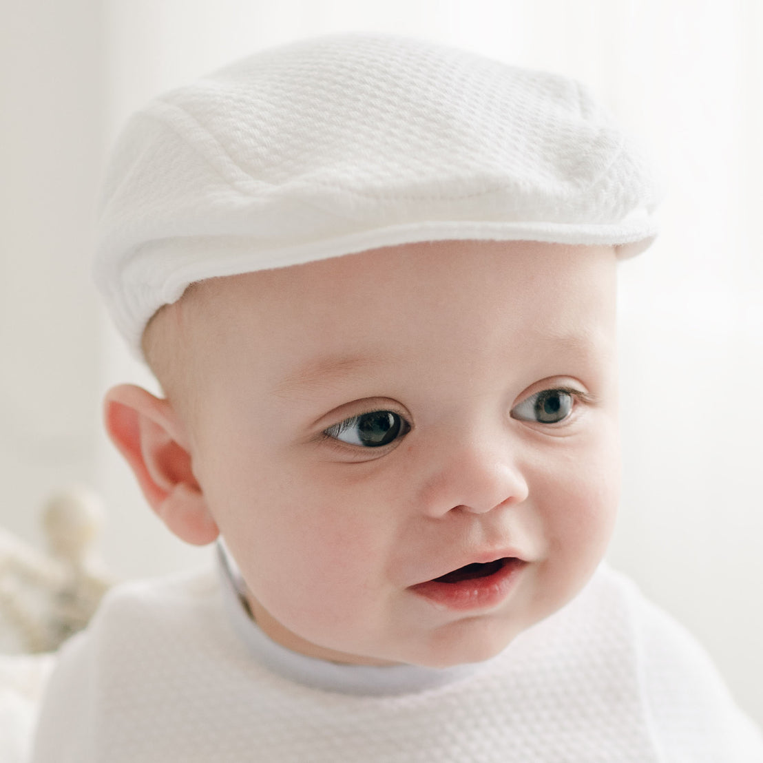 A close-up of a baby wearing the Harrison Textured Newsboy Cap in white, accompanied by a matching outfit. The baby has a curious expression with wide, blue eyes and a slightly open mouth. The background is softly blurred, creating a serene and bright atmosphere.