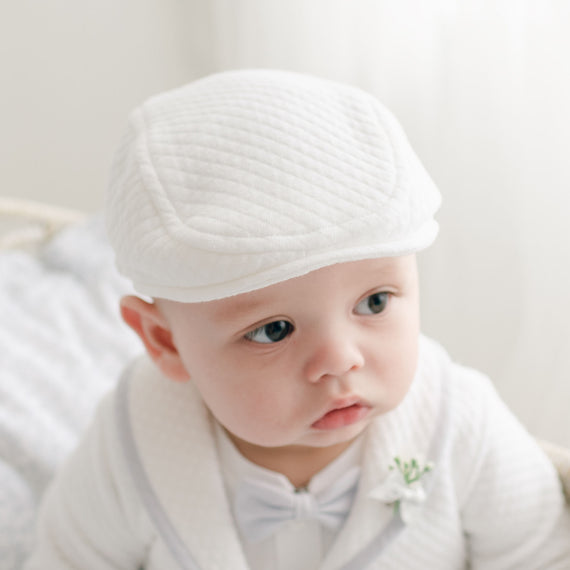 A baby wearing a Harrison Quilted Newsboy Cap and matching outfit with coordinating booties, complemented by a small flower boutonniere, looks off to the side. The background is soft and blurred, drawing attention to the baby's thoughtful expression and delicate features.