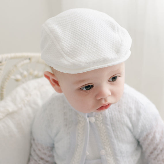 A baby wearing the Harrison Textured Newsboy Cap and a delicate white outfit with intricate detailing sits on a light-colored wicker chair. The baby gazes to the side with a curious expression against a soft, light background. The textured cotton attire is thoughtfully handmade in the USA.