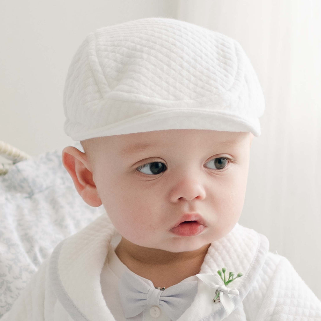 A baby wearing the Harrison Quilted Newsboy Cap and a matching outfit with a small bowtie gazes off to the side. The background is softly blurred, emphasizing the baby's serene and thoughtful expression.