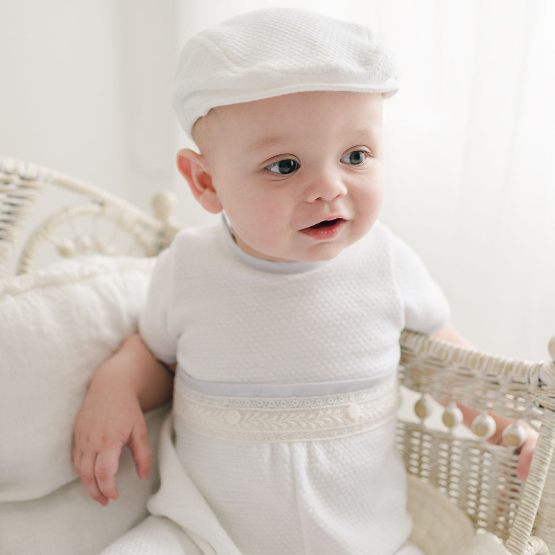 A baby dressed in the Harrison Short Sleeve Romper and a matching cap sits in a wicker chair. The baby is looking slightly to the side with a curious and happy expression. The background is softly lit, enhancing the serene and innocent atmosphere of the scene.