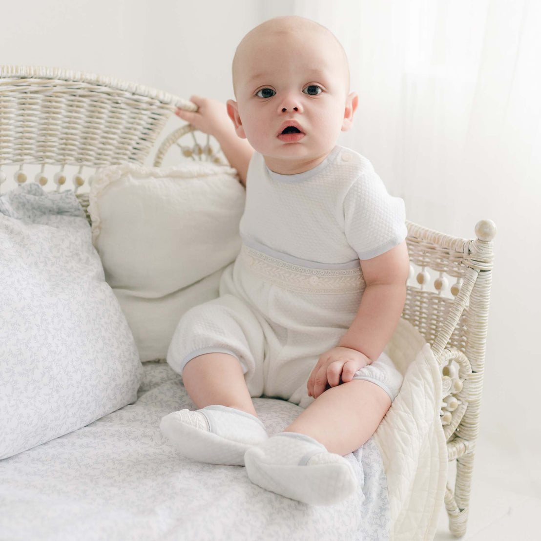 A baby in a Harrison Short Sleeve Romper sits on a white wicker crib, holding onto the side rail. The background is softly lit with white curtains and bedding, creating a serene and peaceful atmosphere. The baby looks towards the camera with a curious expression.