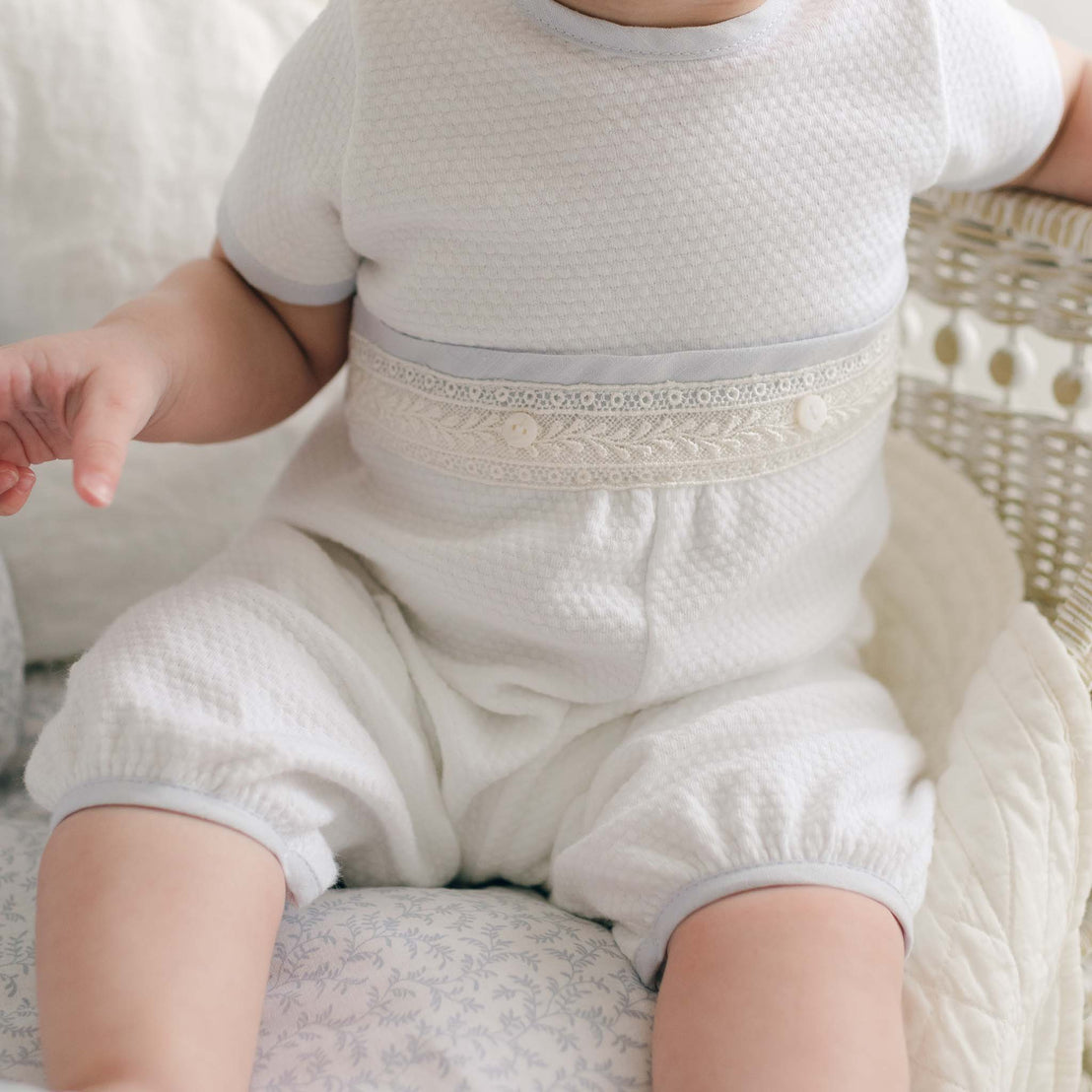 A baby dressed in a Harrison Short Sleeve Romper with decorative lace and button details across the chest sits in a wicker crib. The baby’s face is not visible, and the crib and bedding have a light, neutral color scheme, perfect for a special occasion outfit.