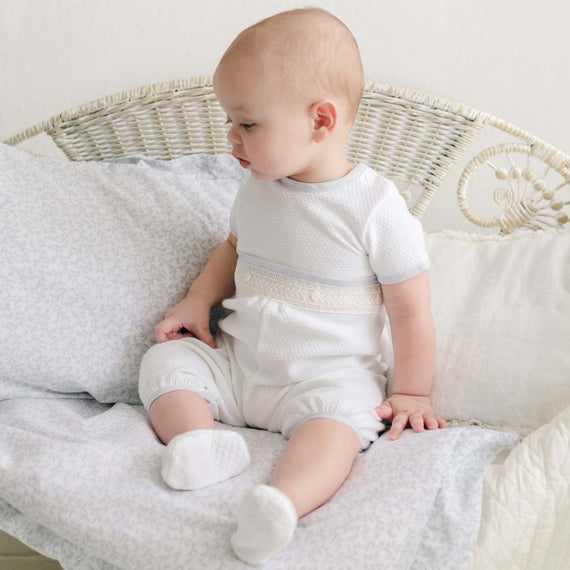 A baby sits on a white wicker bench adorned with a light-colored blanket and cushion. Dressed in the Harrison Short Sleeve Romper, the baby looks to the side, showcasing their adorable special occasion outfit. The plain, light-colored background creates a serene atmosphere.