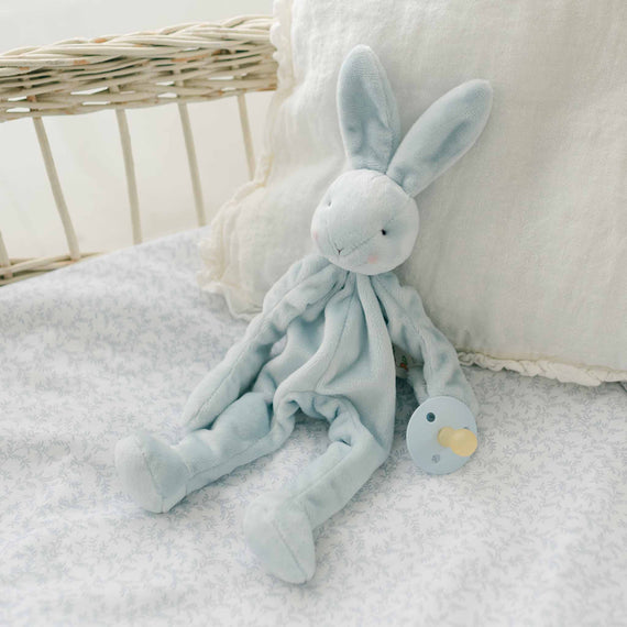 The Harrison Silly Bunny Buddy Pacifier Holder, a soft blue stuffed rabbit toy, serves as a charming pacifier holder on a white and light blue patterned fabric. The rabbit clutches a pacifier in one hand, adding function to its form. Behind it, there's a cream-colored pillow with part of a wicker crib visible—perfect as a thoughtful baby gift.