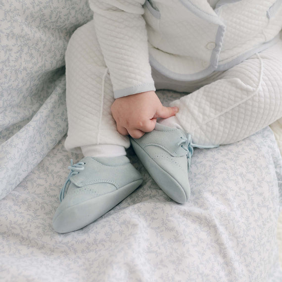 A close-up of a baby wearing a white quilted outfit and Harrison Suede Shoes in soft blue. The baby is seated on a bed with a light floral-patterned sheet, touching one of the handmade in Spain Harrison Suede Shoes. Only the baby's legs and hands are visible.