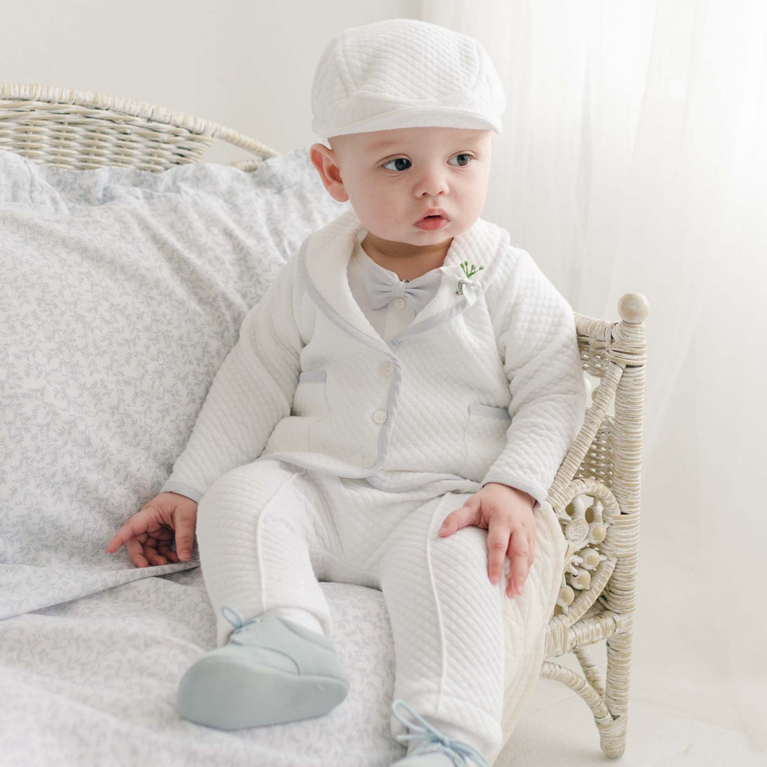 A baby dressed in the Harrison 3-Piece Pants Suit—a stylish white, formal outfit for baptism that includes a hat, jacket, and bow tie—sits on a light-colored cushioned wicker chair. Handmade in the USA, the baby boy suit perfectly complements his curious expression as he looks to the side. The background features soft white curtains and light bedding.