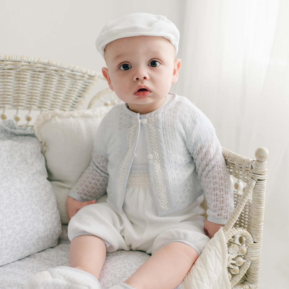 A baby wearing a white hat and the Harrison Blue Knit Sweater sits on a white, wicker daybed with cushions. The baby's curious expression is directed toward the camera. The softly lit background creates a calm and serene atmosphere.